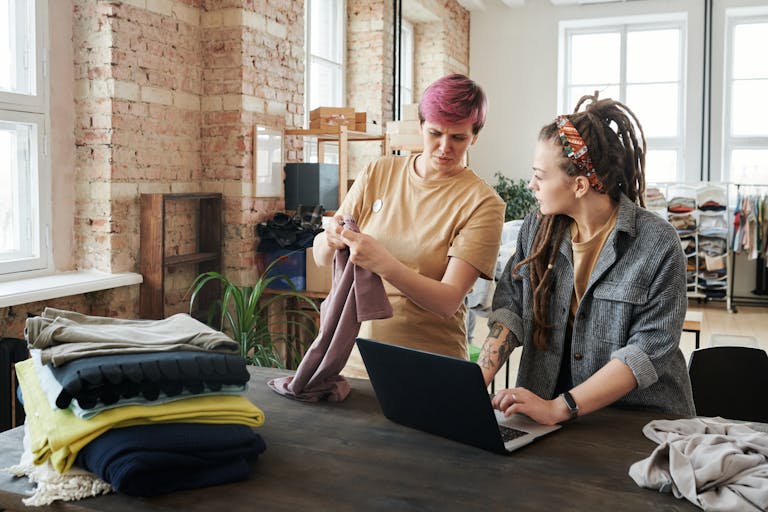 Two women sorting clothing donations using a laptop in a bright urban setting.